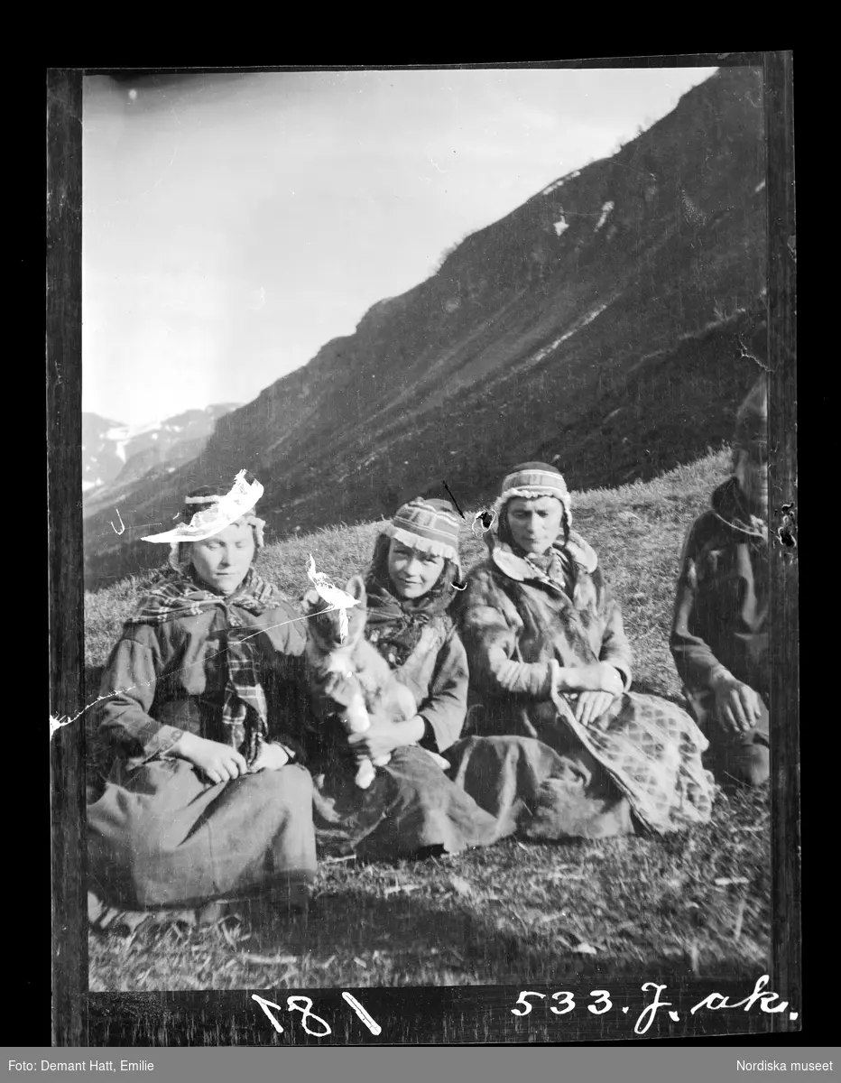 En man, en kvinna (Anne Rasti) och två flickor sitter på marken med ett fjäll i bakgrunden under vårflyttningen från Närvä. Flickan i mitten med en hundvalp i famnen är Marge (Anne och Jouna Rastis dotter). Det är den första vilodagen i Norge efter den mödosamma vandringen över högfjället. Bilden ingår i en serie fotografier tagna av Emilie Demant Hatt i Sapmi mellan åren 1907 och 1916.