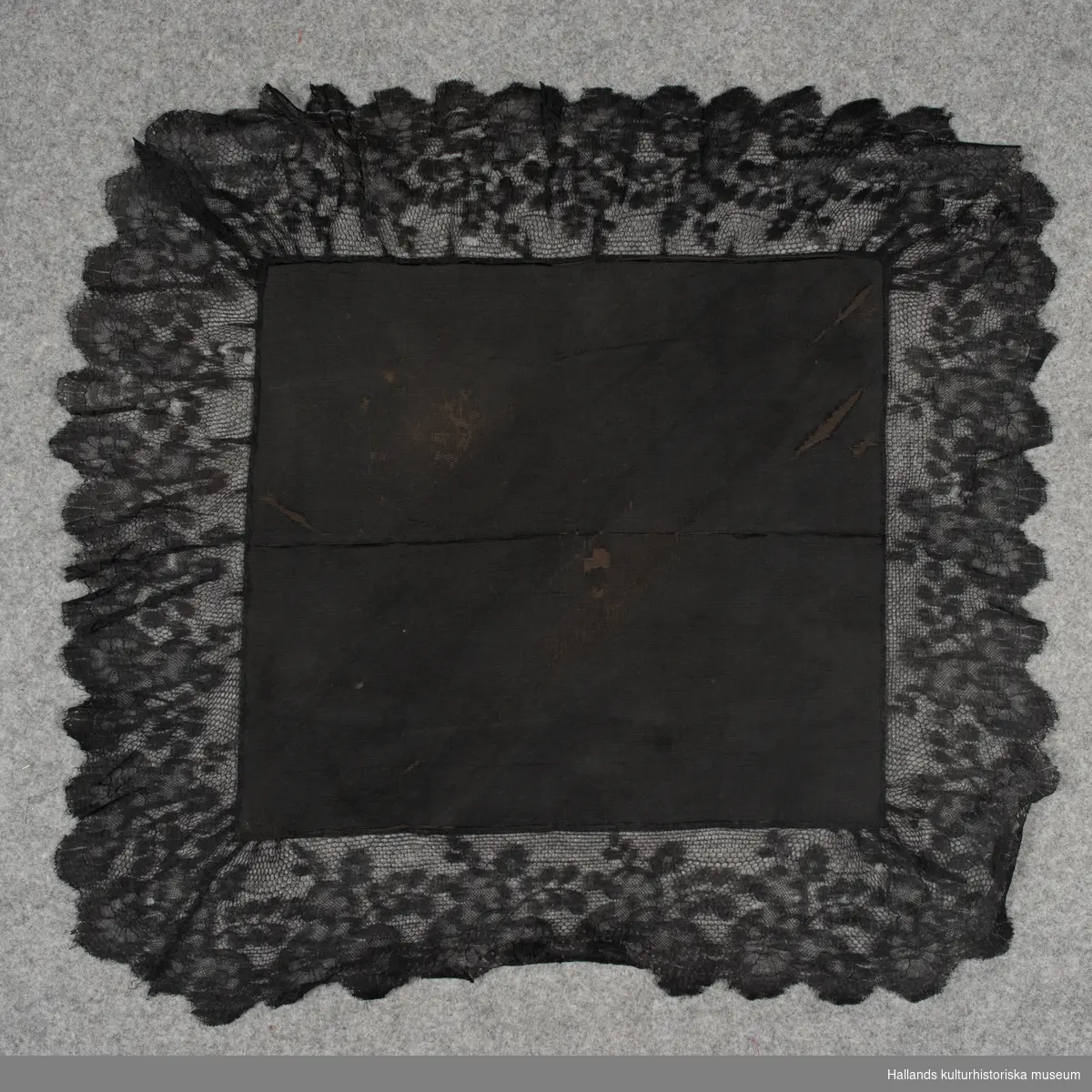 Huvudduk av svart sidenrips, halvsiden (bomull/silke) med bred svart silkespets runt om. Mönstrad med växtranka och bågformad kant.