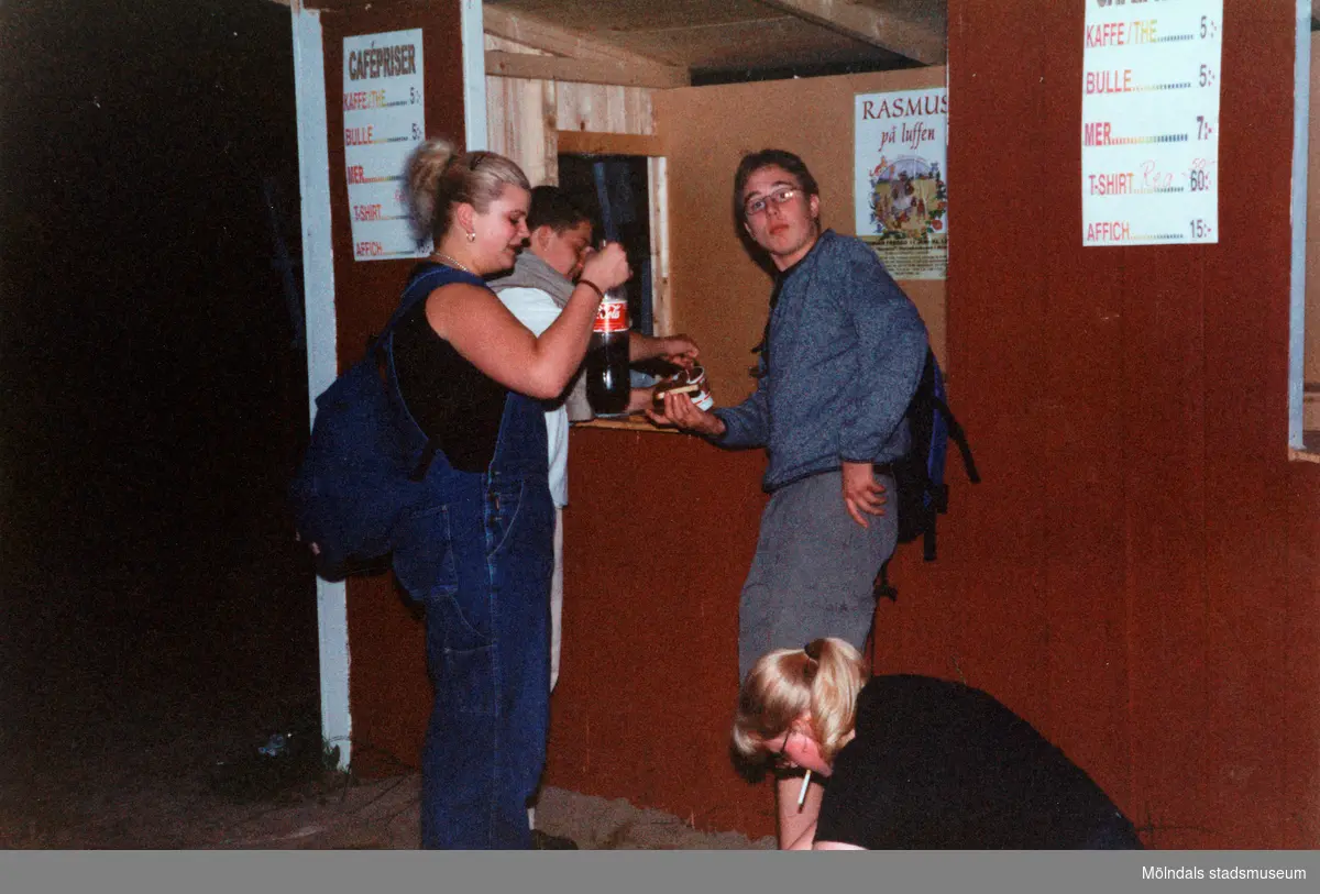Sommarspelet/barnteaterföreställningen "Rasmus på luffen" 1999 vid Byxorna i Gunneboskogen (korsningen Christina Halls väg/John Halls väg). Tre personer (publik) står framför en kiosk. I kiosken står en man och häller upp något.