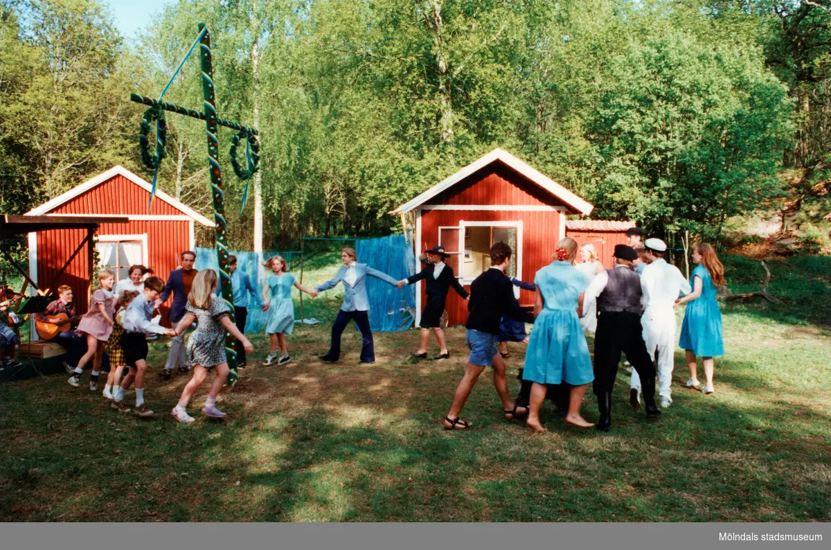 Sommarspelen/barnteaterföreställningen "Tjorven på Saltkråkan" 1996 vid Byxorna i Gunneboskogen. Barn och vuxna dansar runt en midsommarstång. Till vänster ses ett par musiker som spelar på instrument. I bakgrunden ses två små röda stugor som står framför flertalet träd. Relaterade motiv: 2024_0525 - 0533.