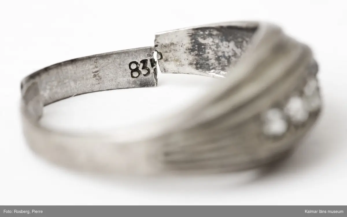 KLM 44496. Ring, fingerring. Av silver med fem slipade stenar på rad. Vita. Själva metallringen i två delar. Stämplar: på utsidan, S och kontrollstämpel, på insidan, 835.