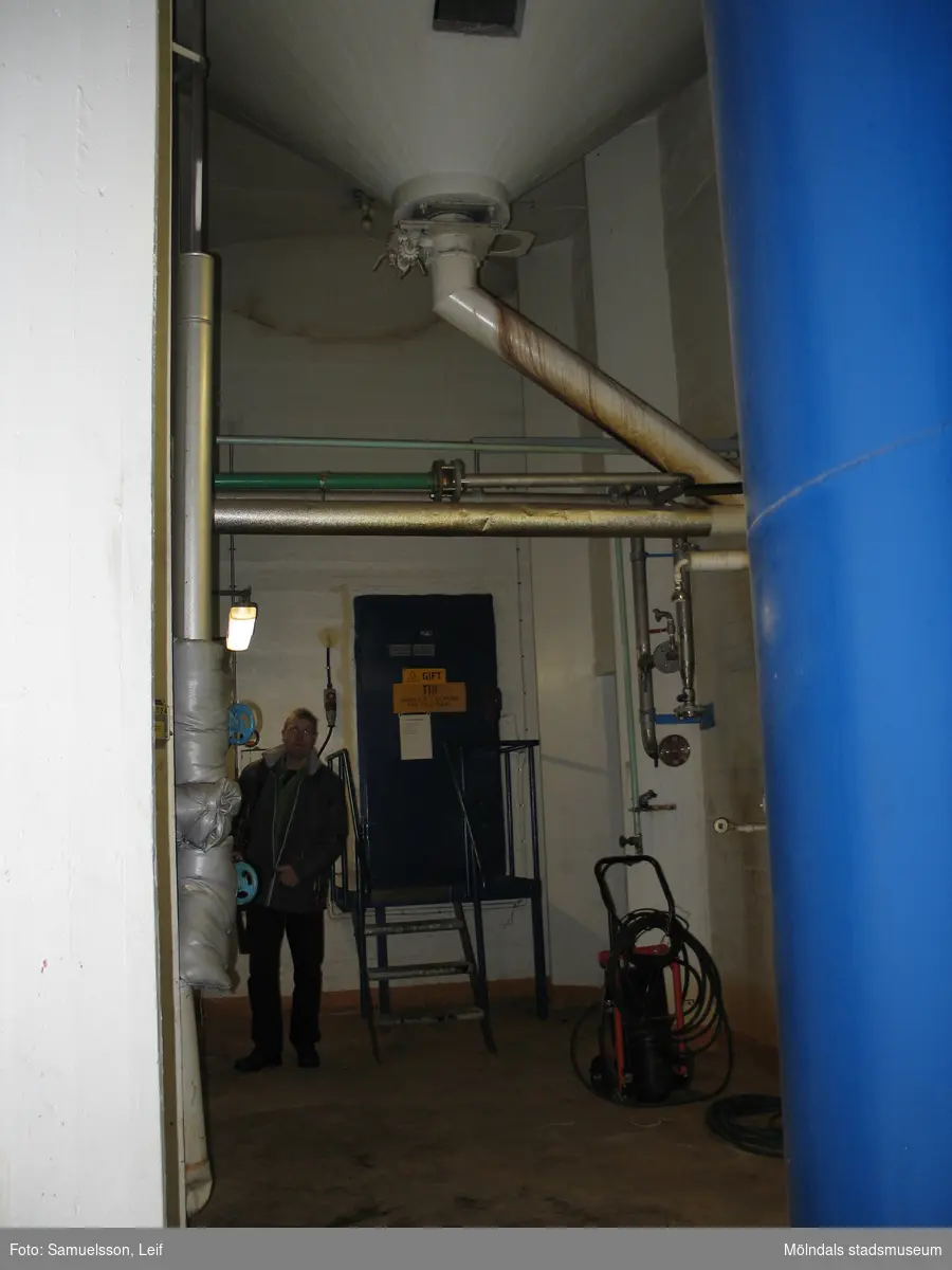 Utrymme med ett utlopp i taket. En man står intill en blå metalldörr med skyltar som varnar för gift. Interiörfotografi från byggnad vid Soabs industrianläggning i Mölndals Kvarnby, år 2007. Anläggningen användes vid fototillfället av Hexion Speciality Chemicals Sweden AB.