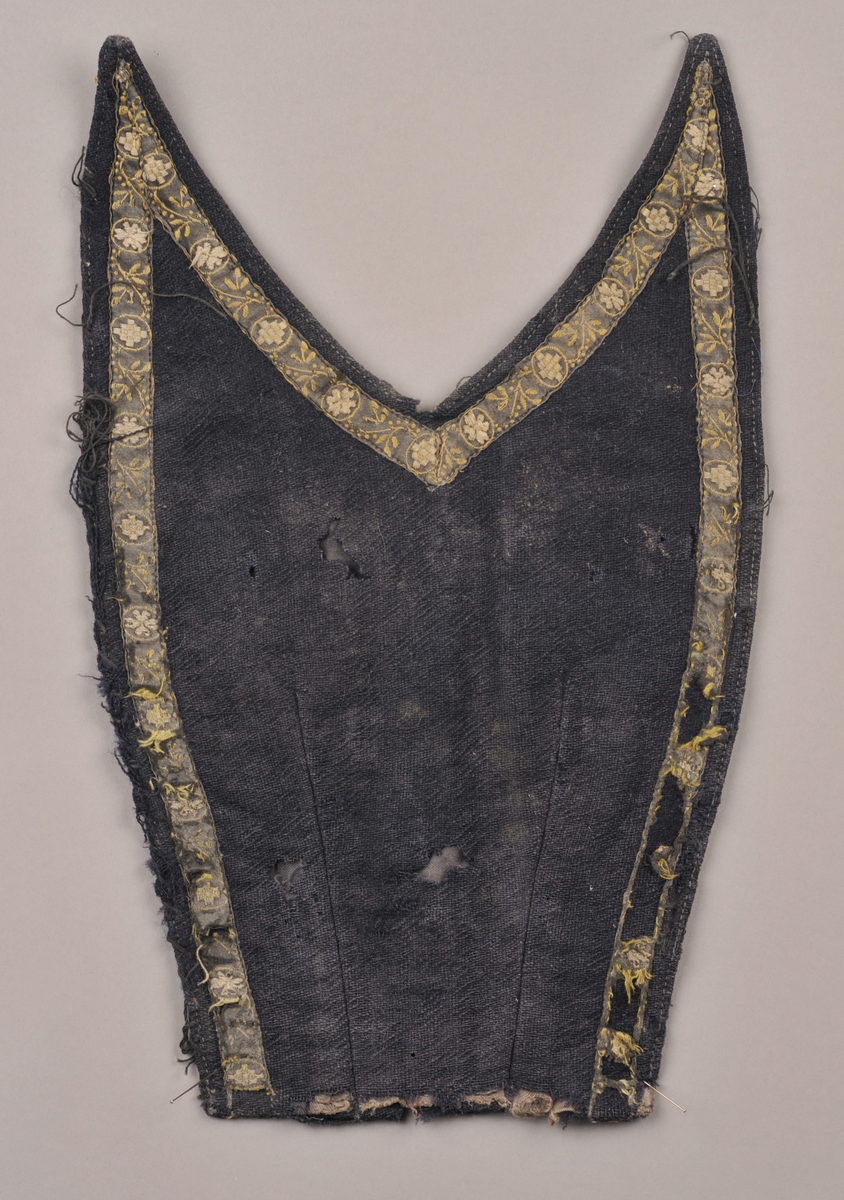 Bringeklut i svart/mørkeblå (?) ulltøy pynta med mønstra band i bomull. Hekter (rusta) på innsida venste side.