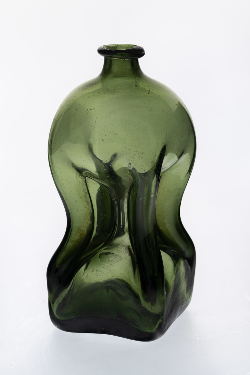 Lav klukkflaske i gjennomskinnelig grønnfarget glass. Timeglass-lignende utforming, hvor den kvadratiske over- og underdelen er forbundet med et tynt rør. Lav hals med glatt kantring, samt puntemerke på undersiden.