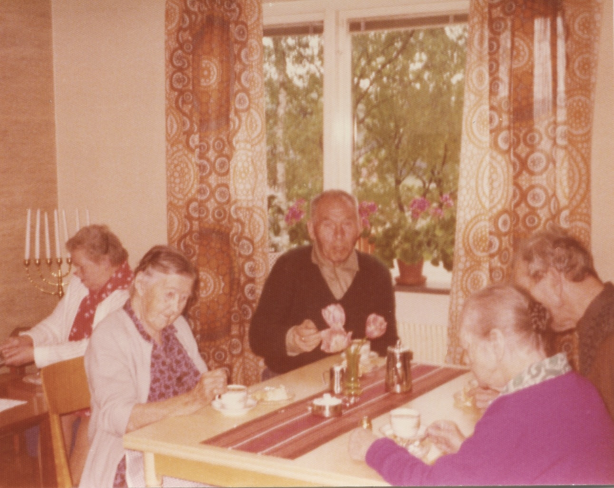 Fikastund för de boende på Brattåshemmet 1970-tal. I bakgrunden sitter en kvinna bredvid en sjuarmad ljusstake. Närmast sitter, från vänster: 1. Selma Börjesson, Högkullen. 2. Okänd man. 3. Okänd kvinna i lila tröja. 4. Okänd man.