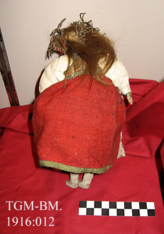 Dukke. Pyntet som brud, på hodet et lad med pynt av messing, rød stakk, rosemønstret forkle. Naturlig hår. Fra protokoll.