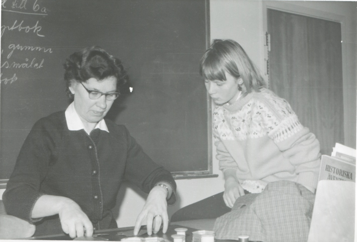 Handarbetslärarinnan Rut Fonell (1903 - 2005) undervisar eleven Kristina Olsson, Brattåsskolan 1965. I bakgrunden skymtar "svarta tavlan".