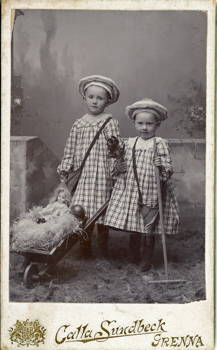 Två små flickor med hatt och rutiga klänningar. De står med liten kratta och skottkärra där det ligge en docka på "halm".