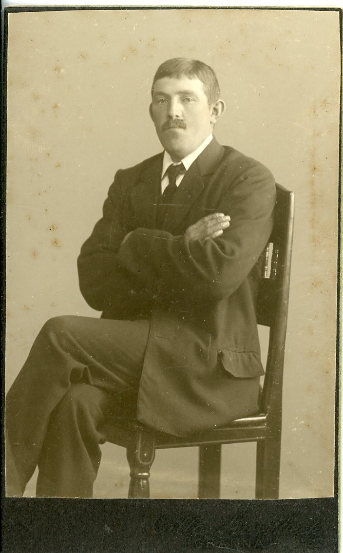 Porträtt av en okänd man med mustasch. Han sitter på en stol med armarna i kors.