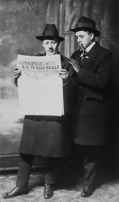 Studiobilde av to menn i hatt, frakk og hansker leser en avis. Overskriften lyder "Australia breaks with U.S. to back Kaiser". Mannen til høyre røyker pipe.