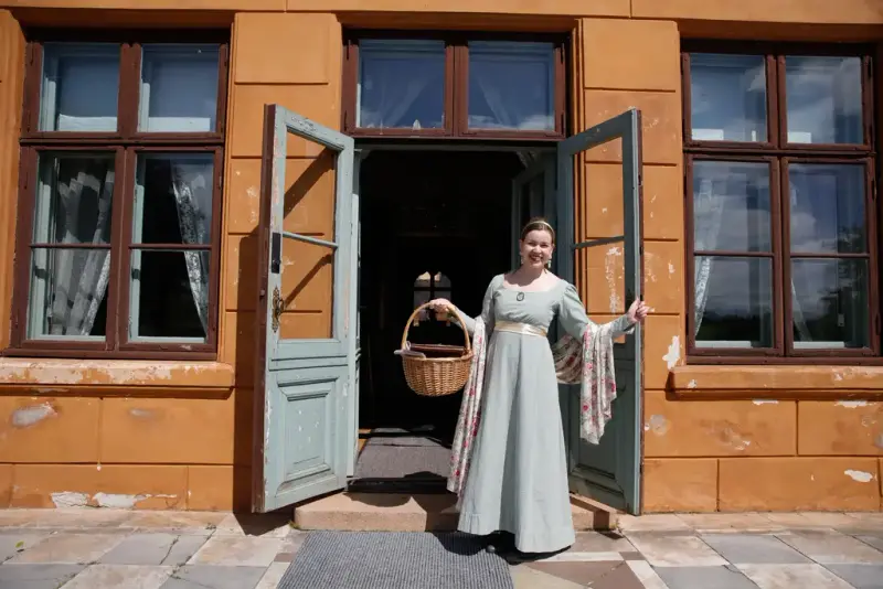 En kvinne i kjole står foran en åpen dør og ønsker velkommen. 