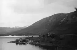 Foto av en eldre fjellgård omgitt av fjord og fjell. Det er 