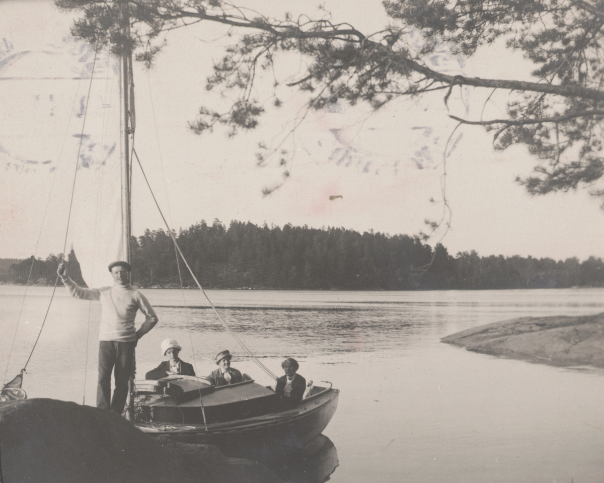 "I Södergarnsviken". Tre kvinnor och man i segelbåt förtöjd vid klippa.