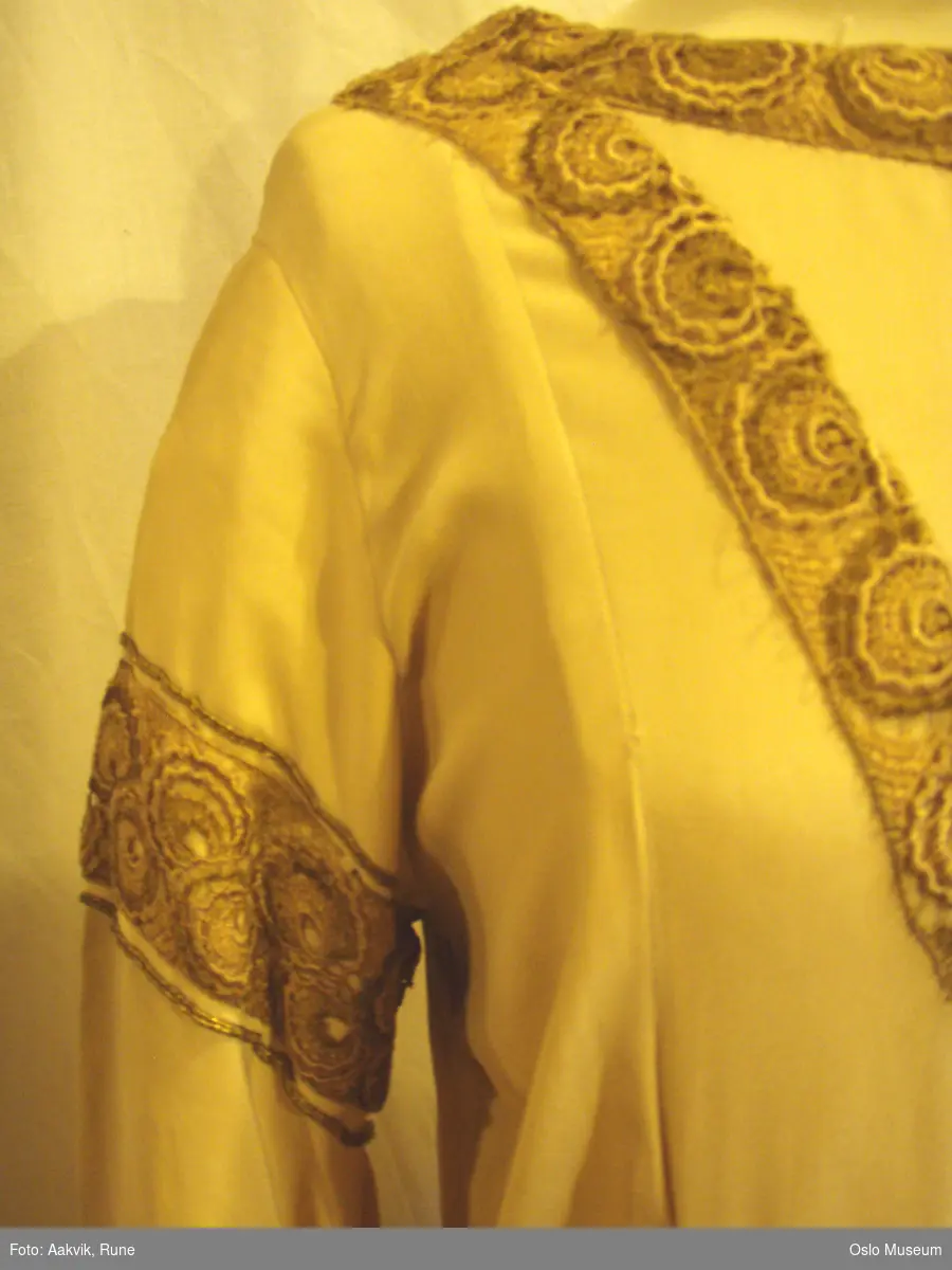 Lang kjole i kremfarget silkedamask med gullfargete dekorbånd. Ermer med slep og belte med gjennombrutt gullfarget spenne.