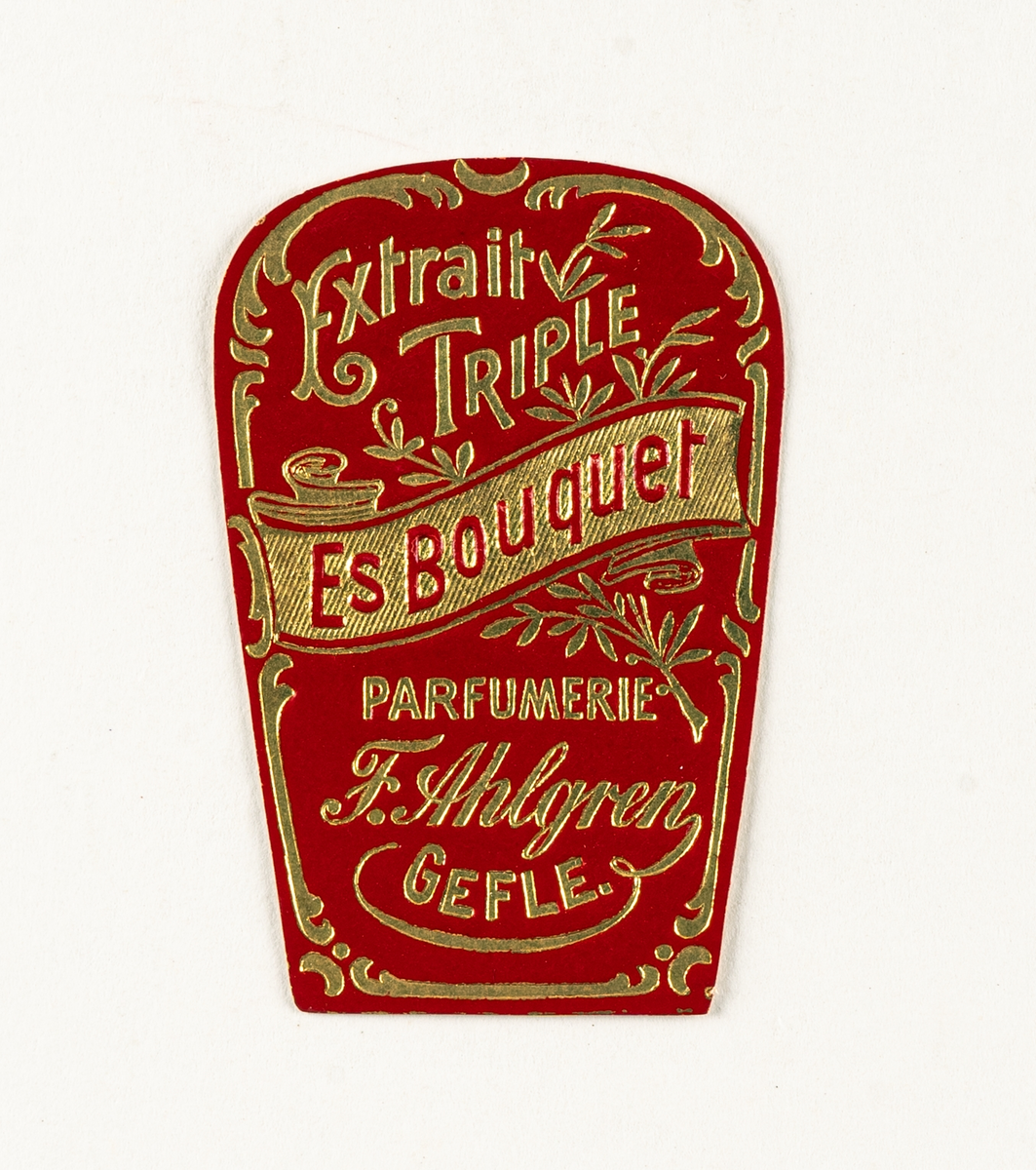 Etikett i papper med blomsterdekor i guld på röd botten med titeln Extrait Triple Es Bouquet Parfumerie F. Ahlgren Gefle.