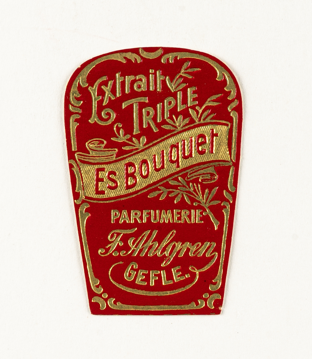 Etikett i papper med blomsterdekor i guld på röd botten med titeln Extrait Triple Es Bouquet Parfumerie F. Ahlgren Gefle.
