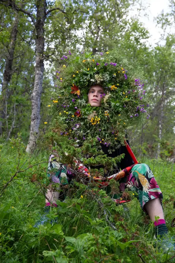 En person sitter på bakken i et skogsmiljø, vedkommende har hyllet seg inn i masse markblomster og kvister. Fargerikt bilde.