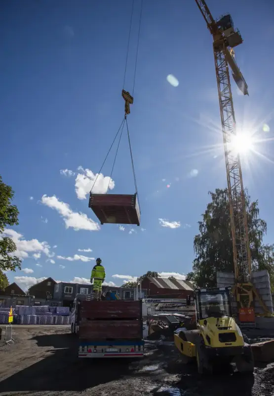 En gul heisekran løfter en stor, firkantet kasse mens sola skinner i bakgrunnen og en mann i signalklær står på et lasteplan klar til å ta imot kassa.