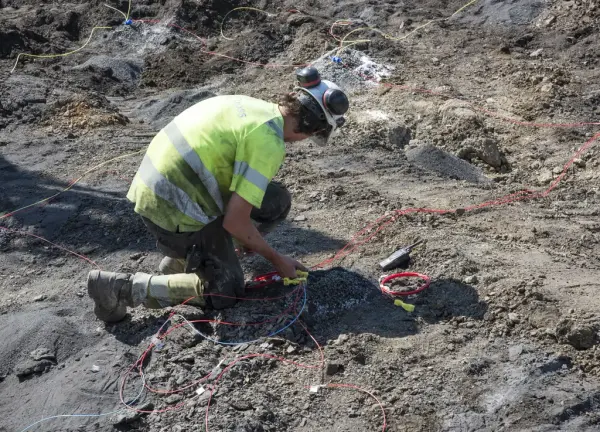 Mann i neongult arbeidstøy står på kne og fester lunte til sprengladning.