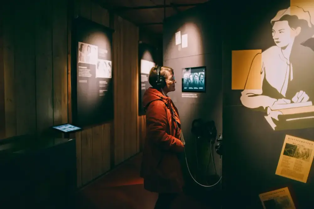 Fotografi i farger tatt i utstillingen om kvinnene på Grini. Midt i bildet står ei kvinne med rød jakke. Hun har på seg høretelefoner og har blikket vendt mot en vegg. Vi ser henne fra siden.