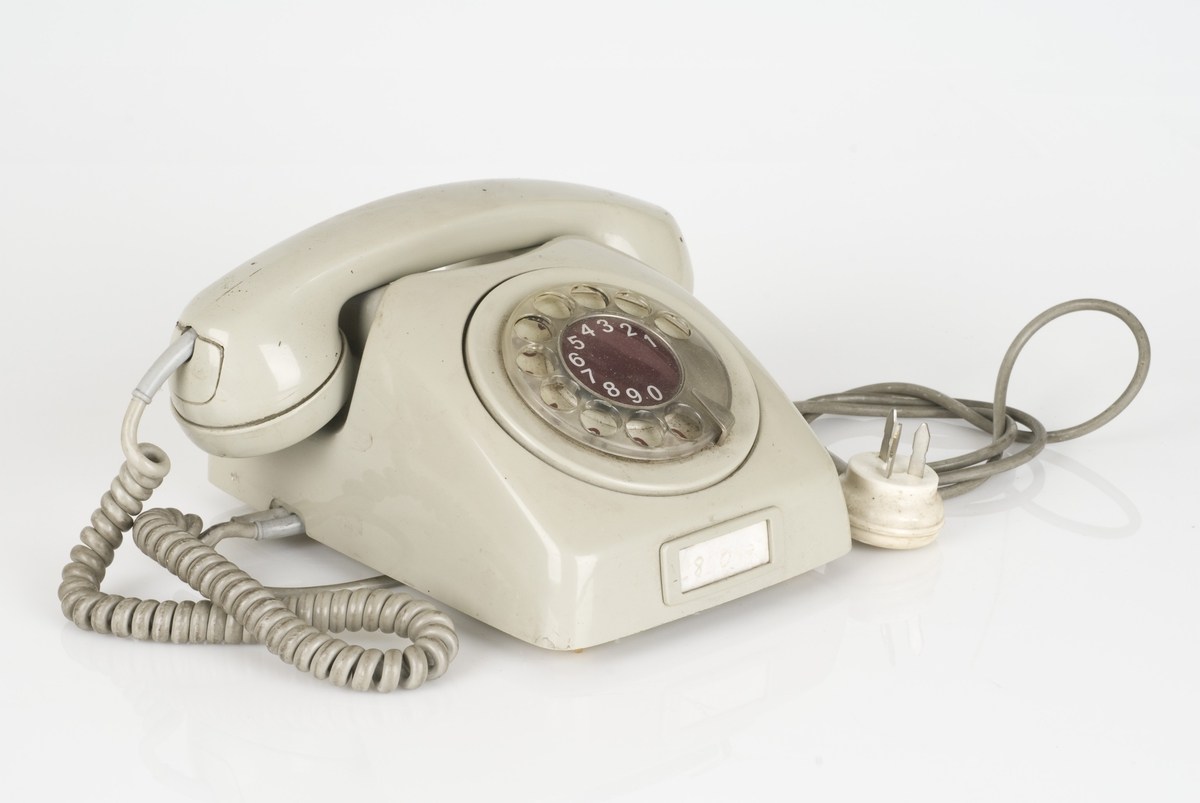 En gammel telefon med roterende tastatur.
Telefonen er av plastog matell. Plasten er i gråfarge.
Påført tekst på undersiden av telefonen.