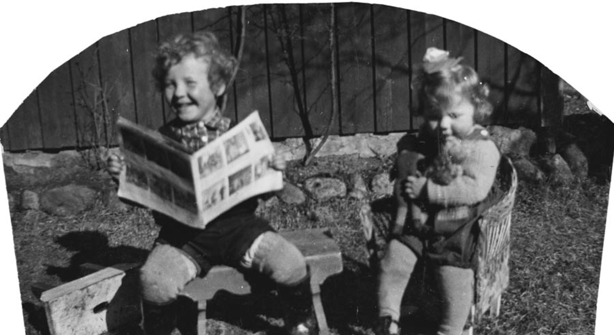 To barn sitter på en skammel og i en barnstol ute og leser tegneserie og koser med bamsen.