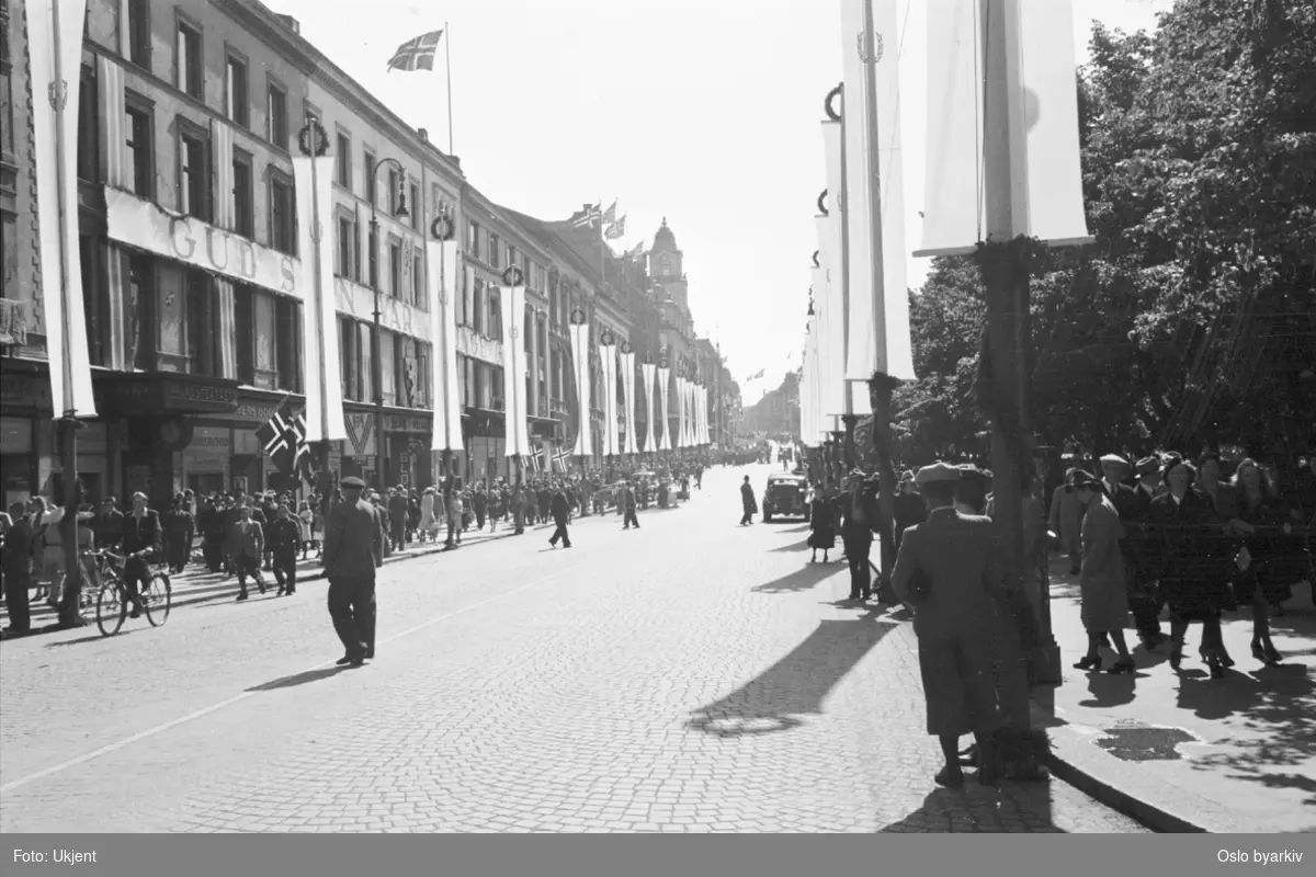 Folkeansamlinger langs en smykket Karl Johans gate 7. juni 1945. (Kongens hjemkomst.) Banner med teksten "GUD SIGN VÅR KONGE GOD" på fasader.