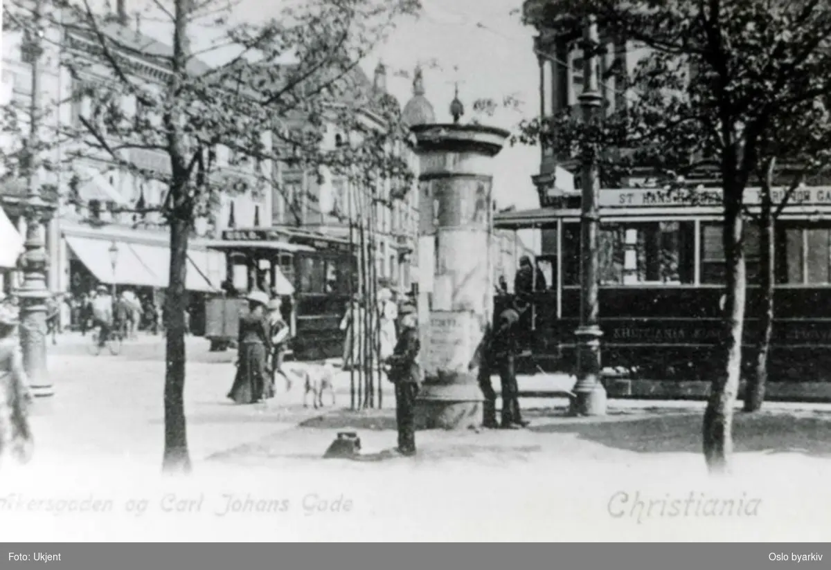 Akersgata krysser Karl Johans gate. Gatemiljø med både hestesporvogn og elektrisk sporvogn. Fra perioden mellom 1894 og 1900. Reklamesøyle.