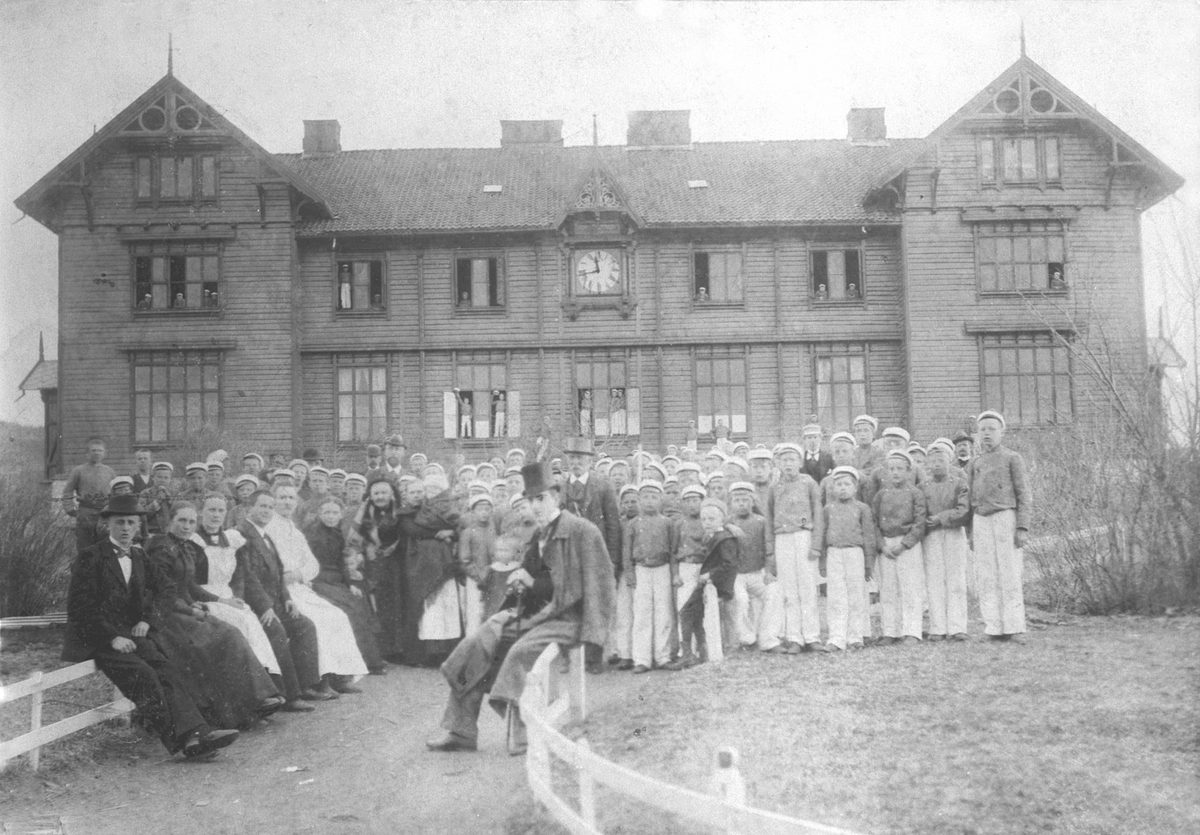Stor gruppe lærere, betjening foran hovedbygningen på Toftes Gave, Nedre Sund, Helgøya. Noen av guttene i uniformen til korpset. Navneliste foreligger på registreringsskjema.