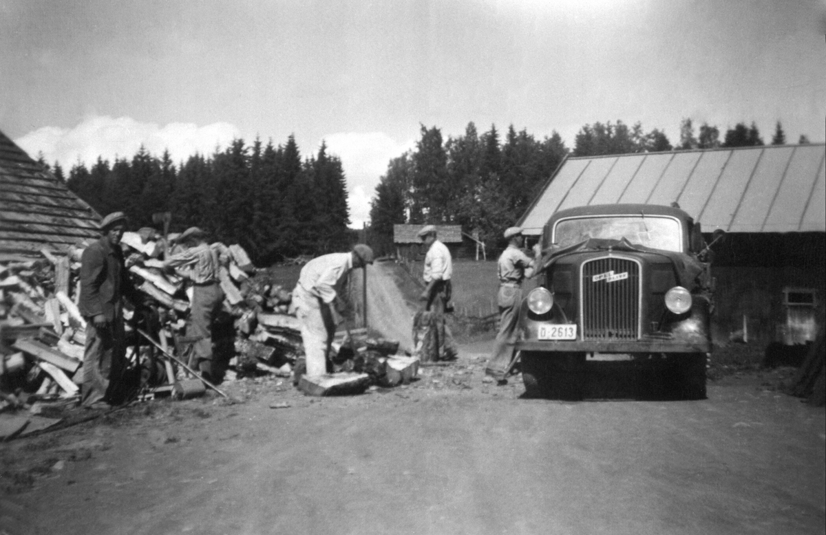 Vedkløyving på Helseth, Stavsjø, Hedmark. Lastebilen er en Opel Blitz med nummer D-2613. I bakgrunn er den gamle smia og gardsvegen. Fra venstre er John Hagelund, Karl Helseth (1919-2002), Ole Nygård, Johan Melby, Anders Helseth (1912-1979).