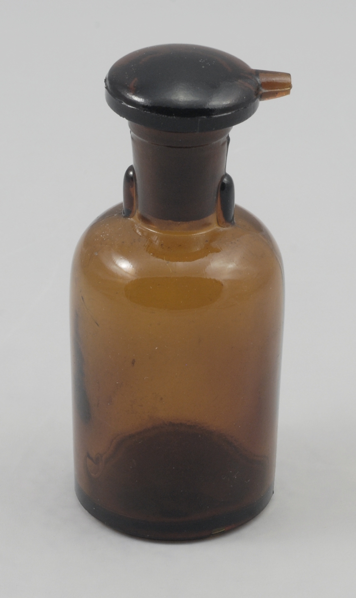 Sylindrisk brun glassflaske med markert avrundet skulder, sylindrisk, lettere konisk hals. På toppen en kork med soppform, korken har rift i stammen og hatten til uthelling av væske.