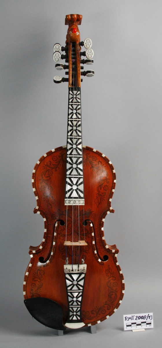 Instrument med fire strenger, og fire understrenger. Rikt dekorert. Halsen ender i et utskåret hode.