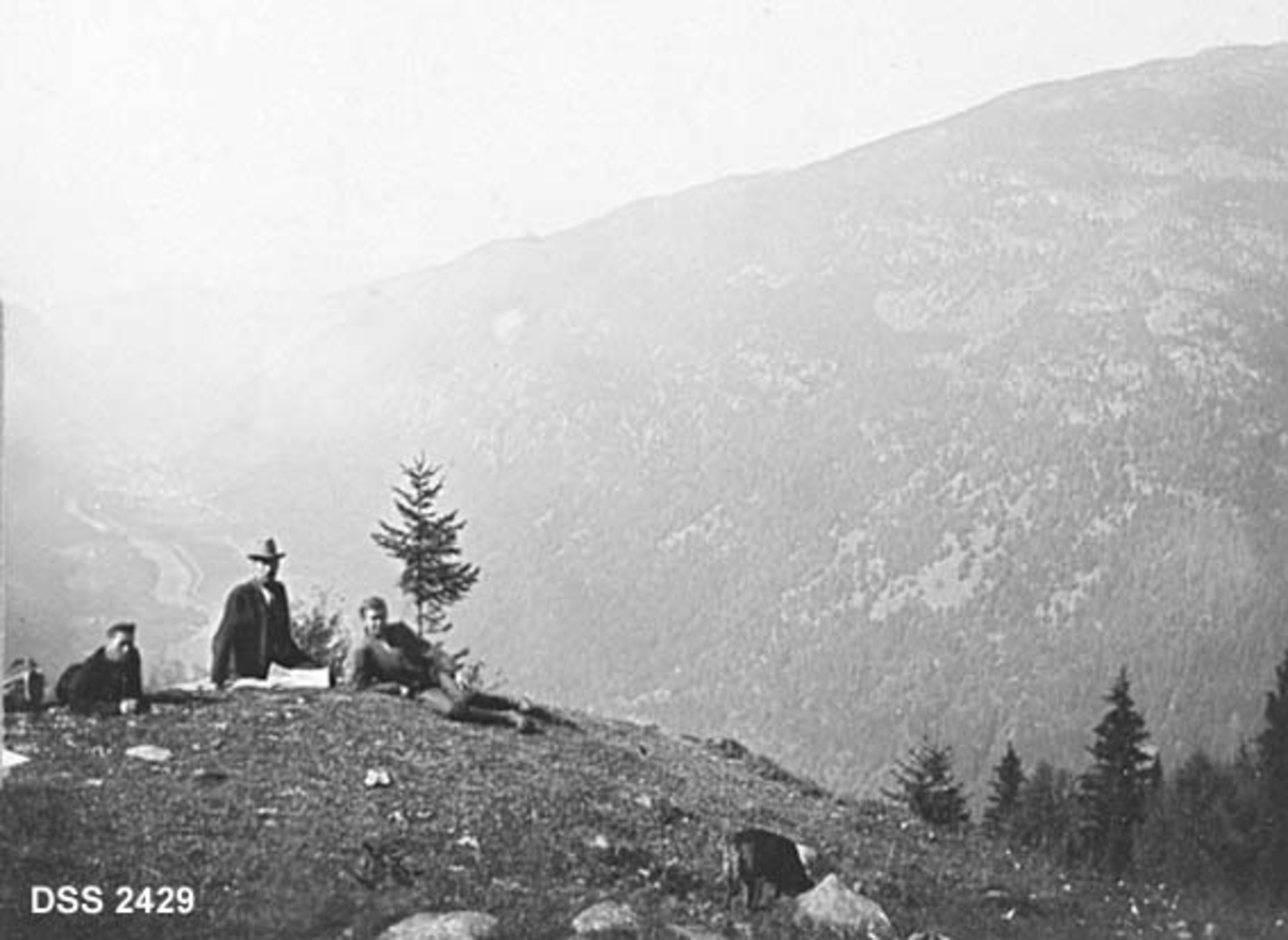Utsiktsbilde fra Tinn.  Bildet er tatt på en grasbevokst haug, der det sitter og ligger tre mann som er på grenseoppgang.  I bakgrunnen til høgre ei fjellside med spredt skog og bak mennene Vestfjorddalen med industristedet Rjukan. 