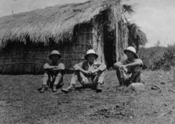 Tre menn sitter på bakken utenfor en strå-/bambushytte i Uga