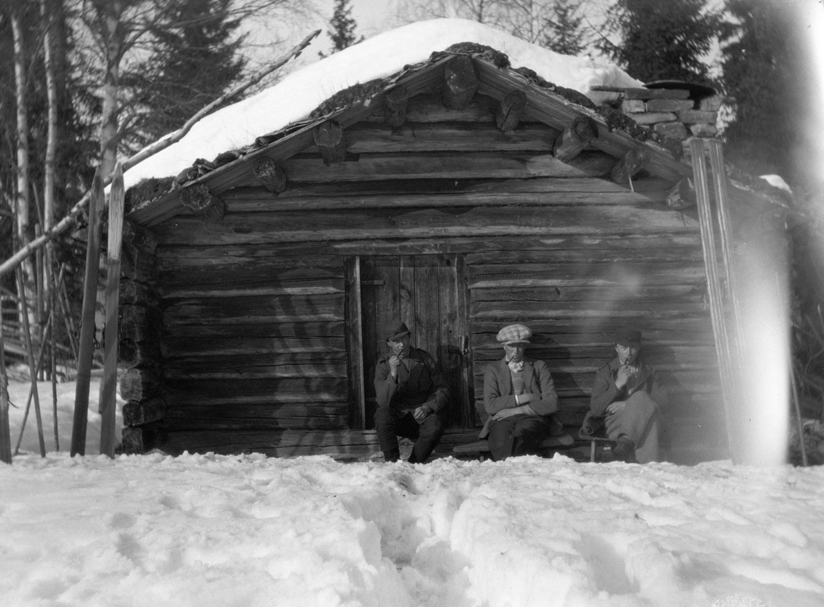 Eksteriør, skog, ski, vinter. Tre ukjente menn røyker pipe ved ei koie. 