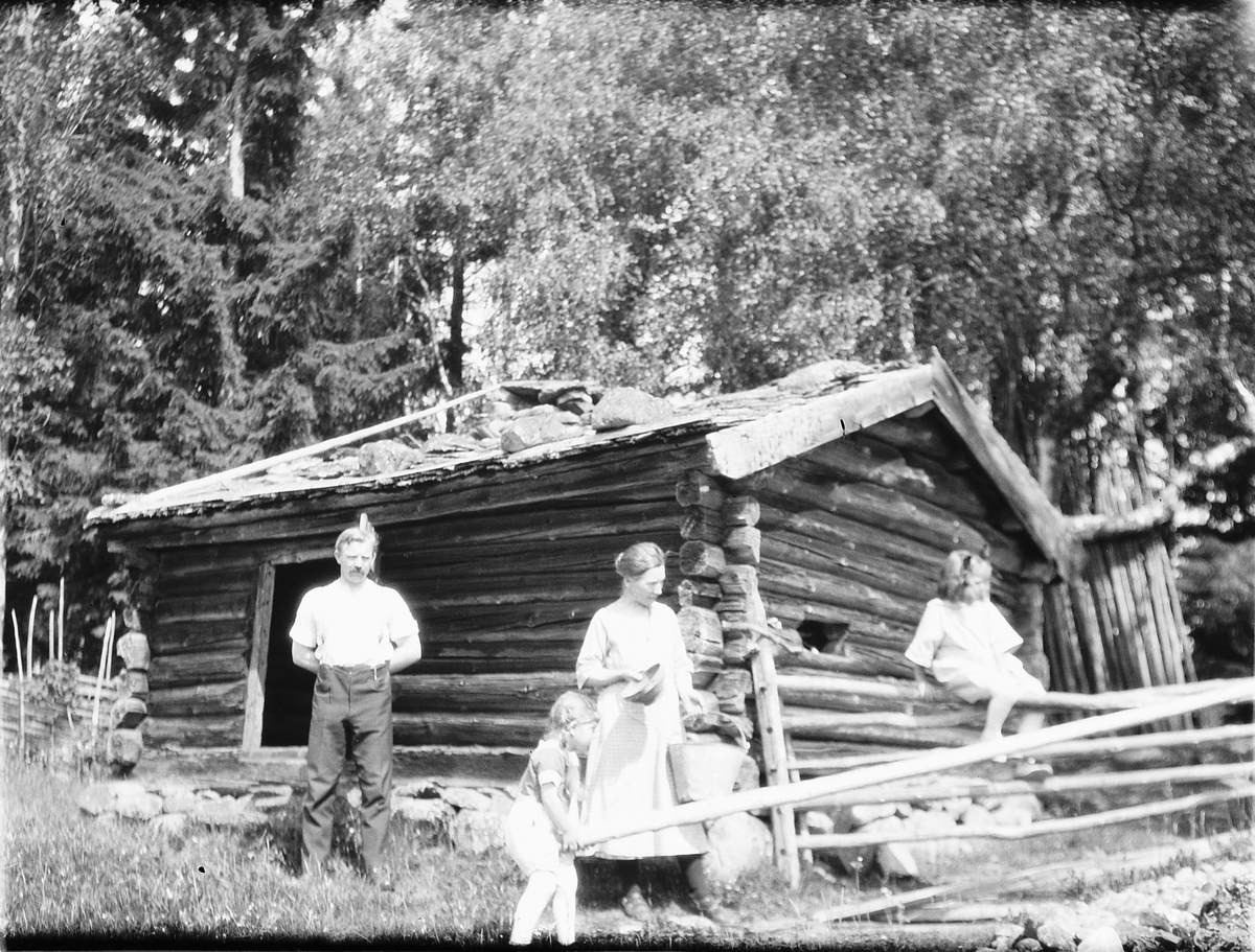 Ukjent mann, kvinne og to jenter ved ei tømmerhytte. 