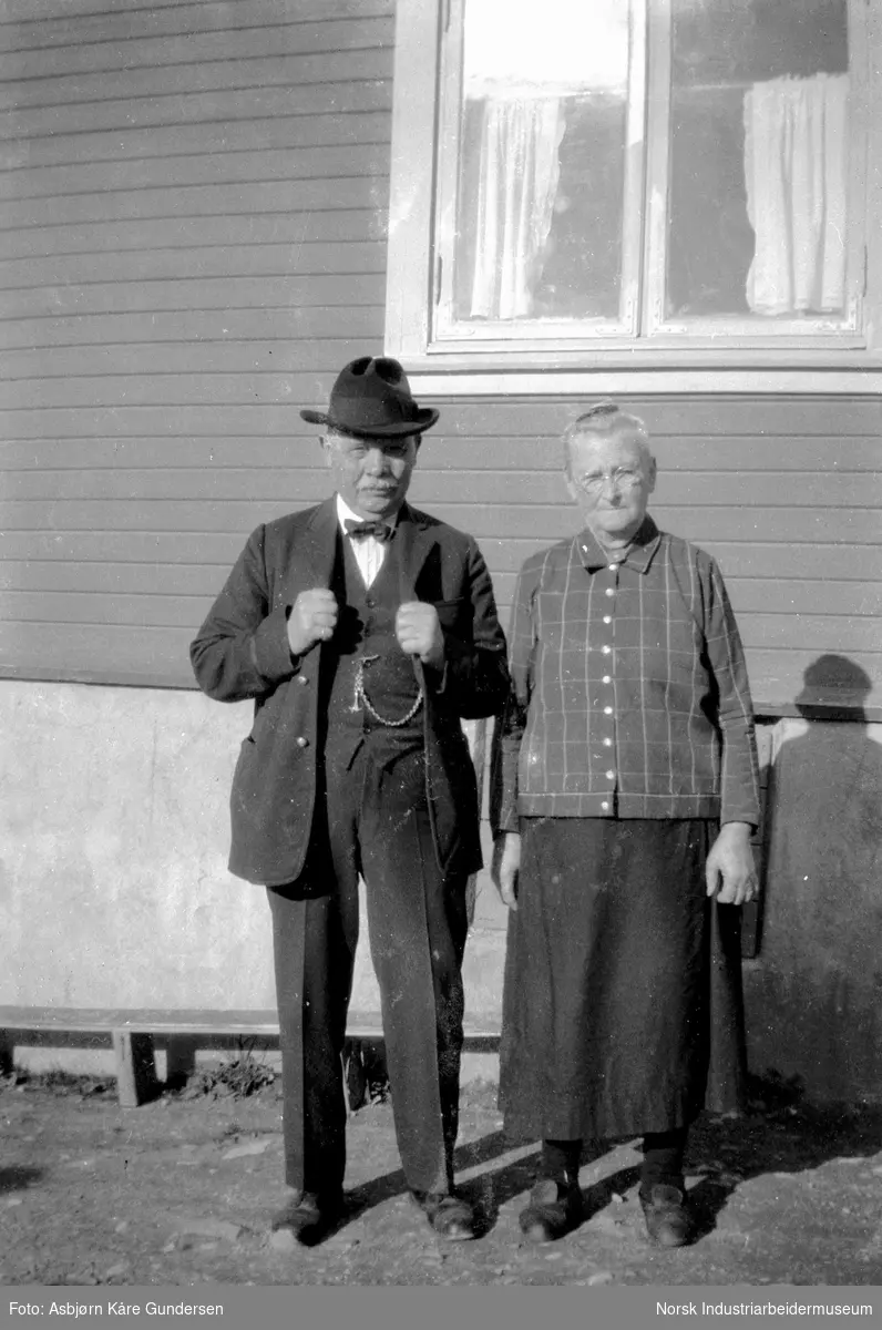 To eldre personer foran en husvegg
