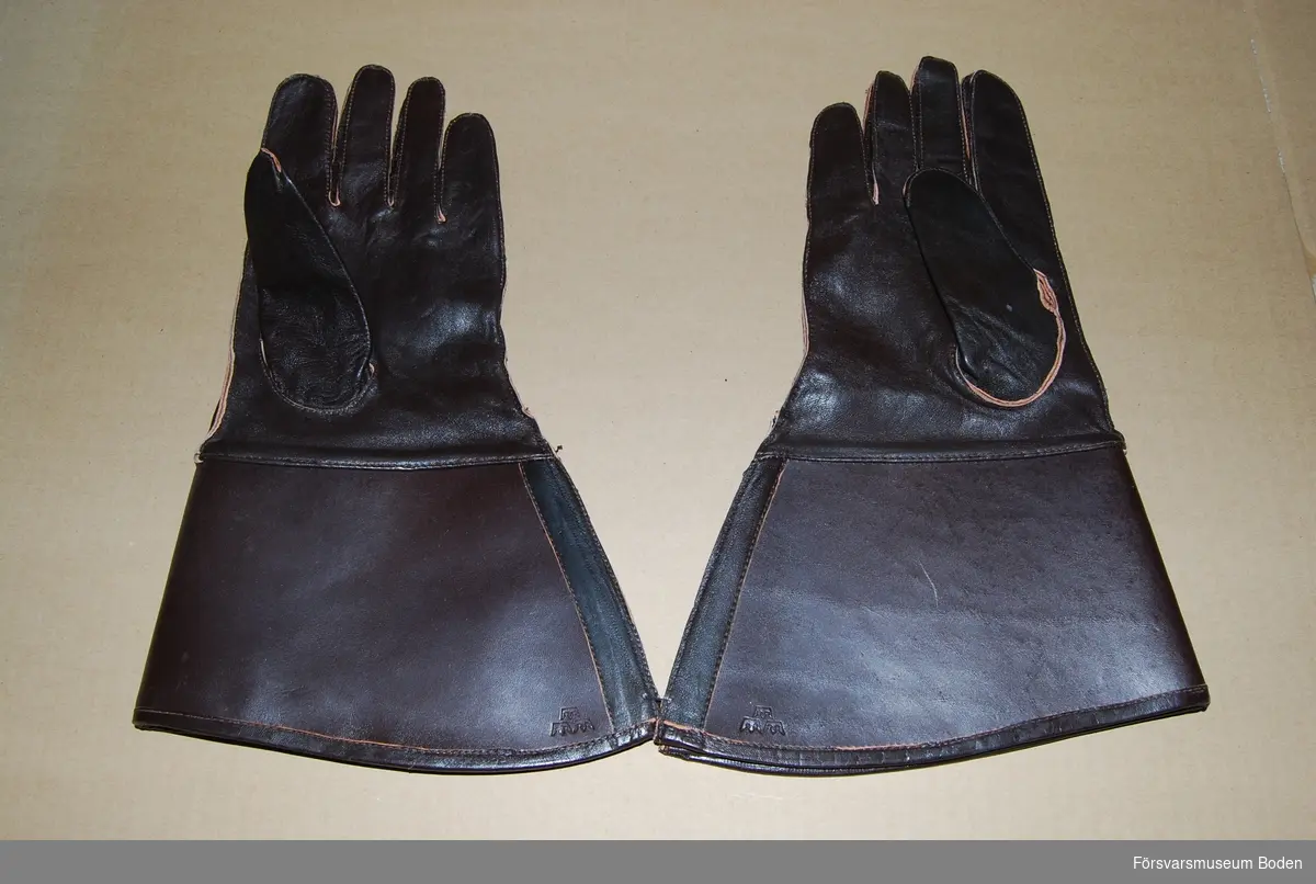 Ett par oanvända handskar av mörkbrunt läder med styv krage som är präglad med tre kronor vid kanten på utsidan. Spänntamp med tryckknapp på ovansidan. Handskarna är hopfästa med tråd. Avsedd att användas av bl.a. mc-ordonnanser.