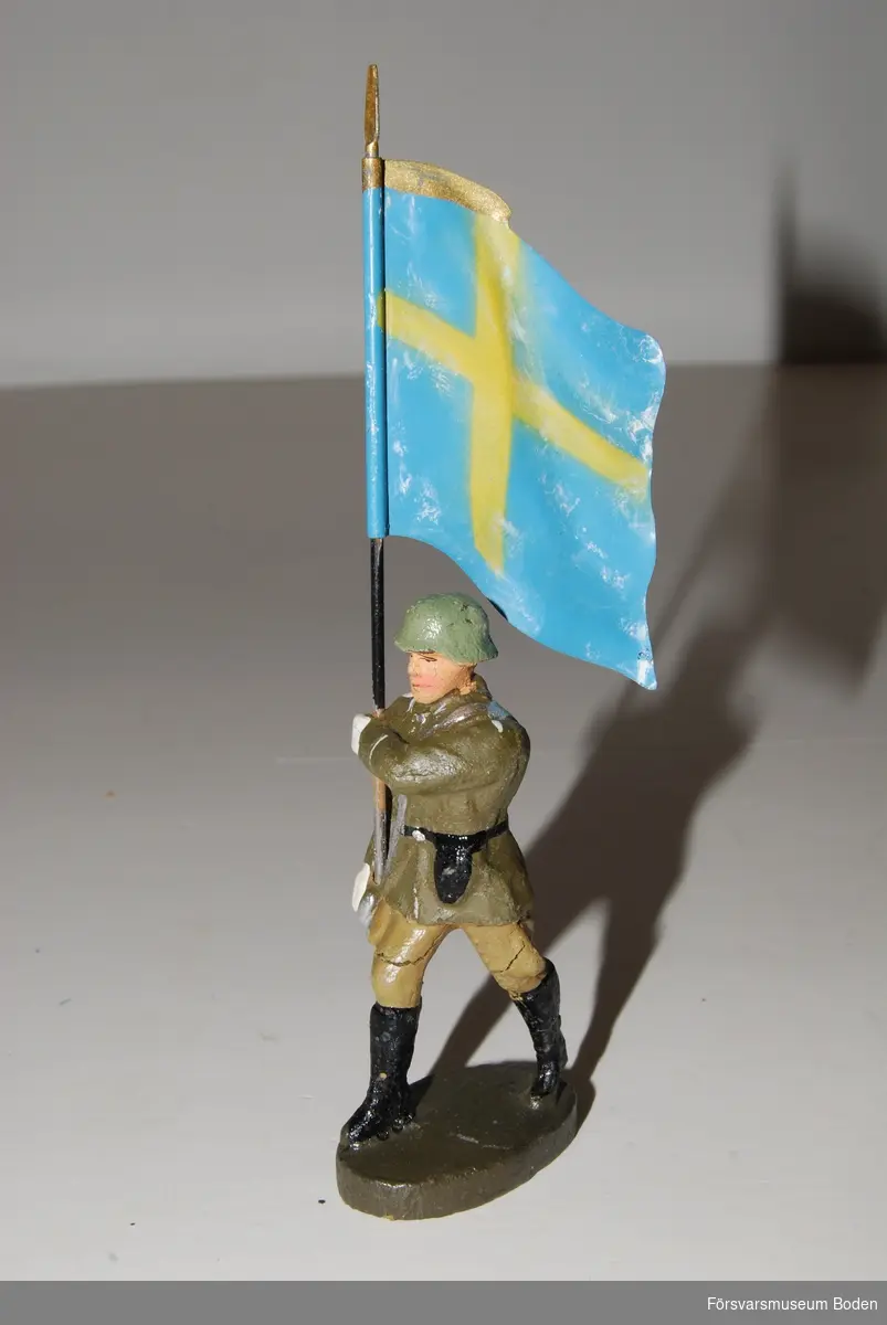 Marscherande fanbärare. Av gjutmassa med flagga i bleckplåt. Tillverkad i Tyskland före 1943. Kan vara konverterad i Sverige innan försäljningen.