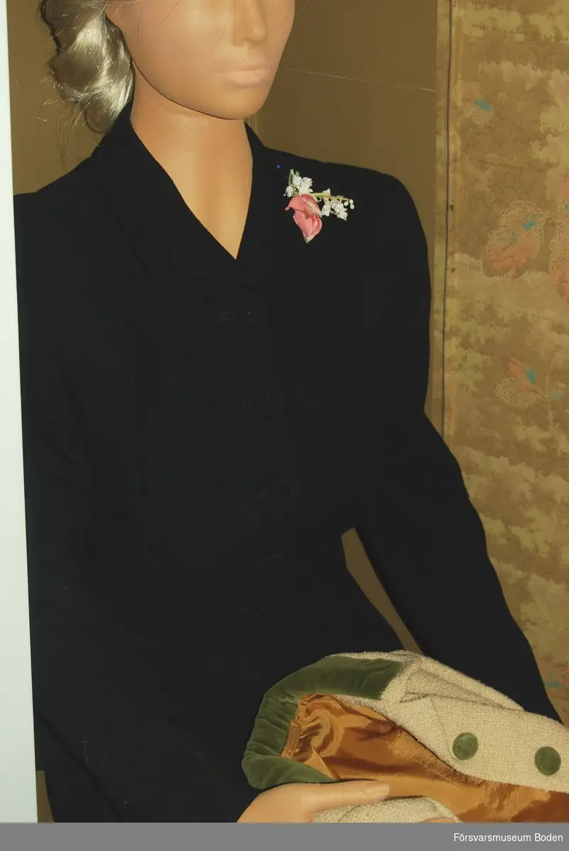1930-talsdräkt i svart ylle. Figursydd fodrad kavaj med klädda knappar och inbyggda axelvaddar. Veckad ofodrad kjol med blixtlås i sidan. Brosch med ros och liljekonvalj som använts till dräkten sitter monterad under vänstra kavajslaget.