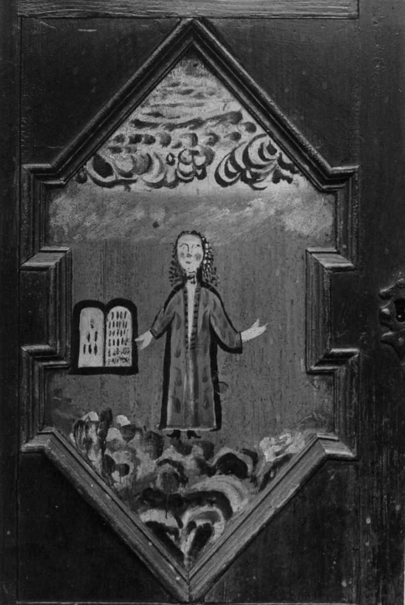 Skap, framskap i en del, to dører oppe og nede. Bibelske motiver; Moses og de ti bud. Gud på en sky, en brennede sky eller busk, en mann som har mistet en sko og en stokk.