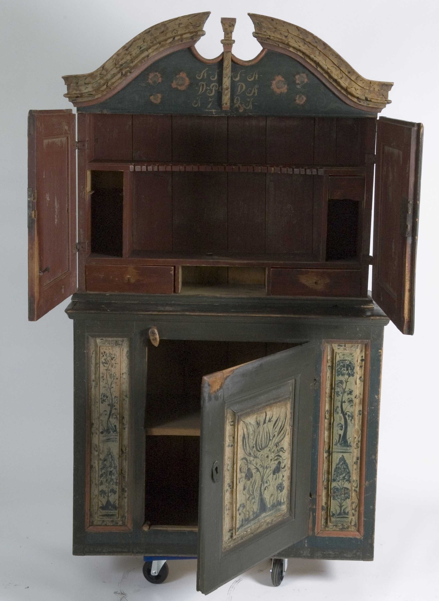 Framskap i to deler. Buet topplist. Skuffer og skjehylle i overskap. Malt innvendig og utvendig. Kirke og bygning malt på dør på overskap. Planter på underdel. Marmorert på lister på overskap.