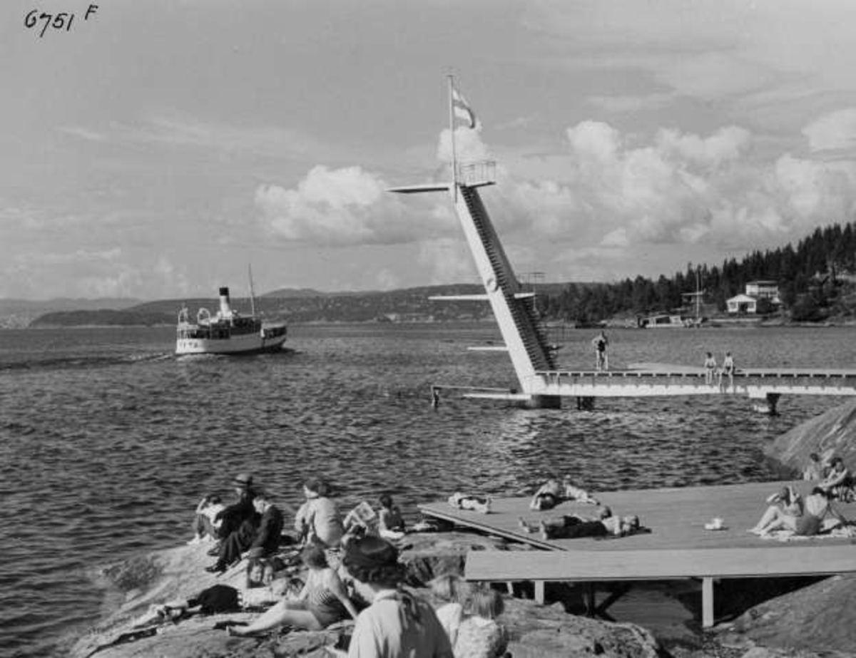 Strandliv med badegjester som soler seg på svaberget ved sjøen på Ingierstrand bad utenfor Oslo. Stupetårn og båt i bakgrunnen.
