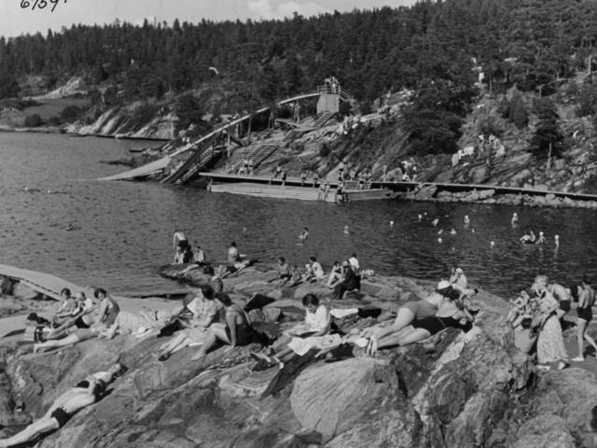 Strandliv med badegjester som soler seg på svaberget ved sjøen på Ingierstrand bad utenfor Oslo. Vannsklie i bakgrunnen.