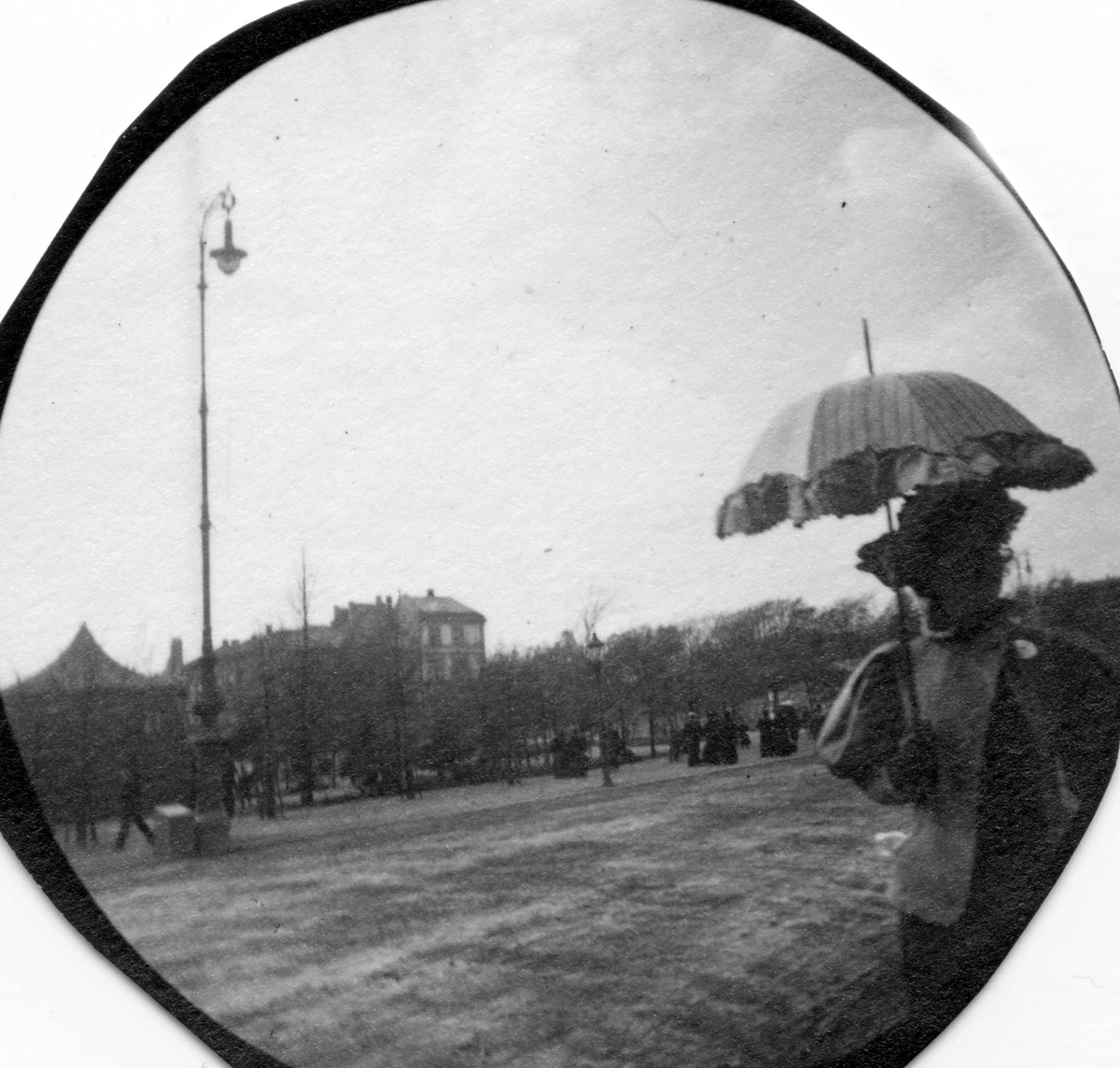 Kvinne med parasoll spaserer i park, Oslo. Bygårder i bakgrunnen.