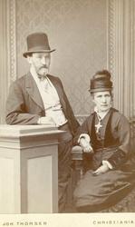 Portrett. Eldre par, mann står og kvinne sitter i fotoatelie