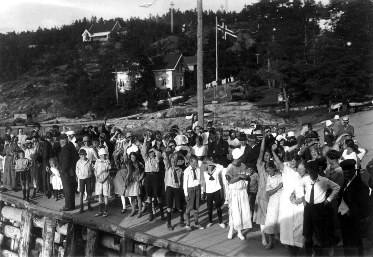 Hvitsten -  Vestby, Akershus 1924. Oversiktsbilde. Festkledde mennesker samlet på brygge. Hus i bakgrunnen og flagg.