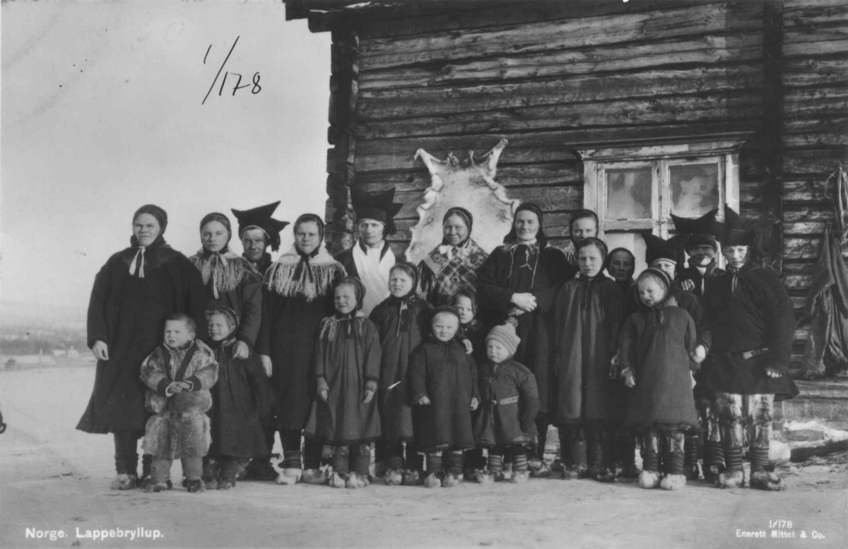 Brudepar og bryllupsgjester, barn og voksne, foran en husvegg med skinnfell, antagelig i Karasjok.