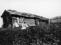 Kvinne i folkedrakt sittende utenfor våningshus, ukjent sted