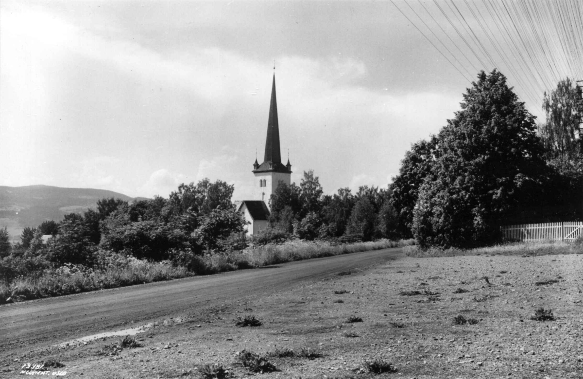 Ringsaker kirke, eksteriør, juni 1935. Omgitt av trær. Vei i forgrunnen. Åser i bakgrunnen.